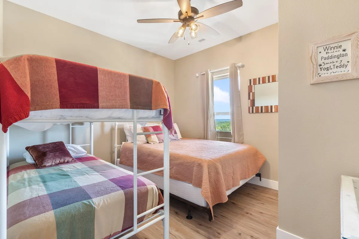 A cozy bedroom featuring a twin-over-twin bunk bed and a queen bed, situated alongside a window that frames a lovely outdoor view. The space is styled with soft bedding and gentle, neutral colors to promote relaxation and comfort for both adults and children.