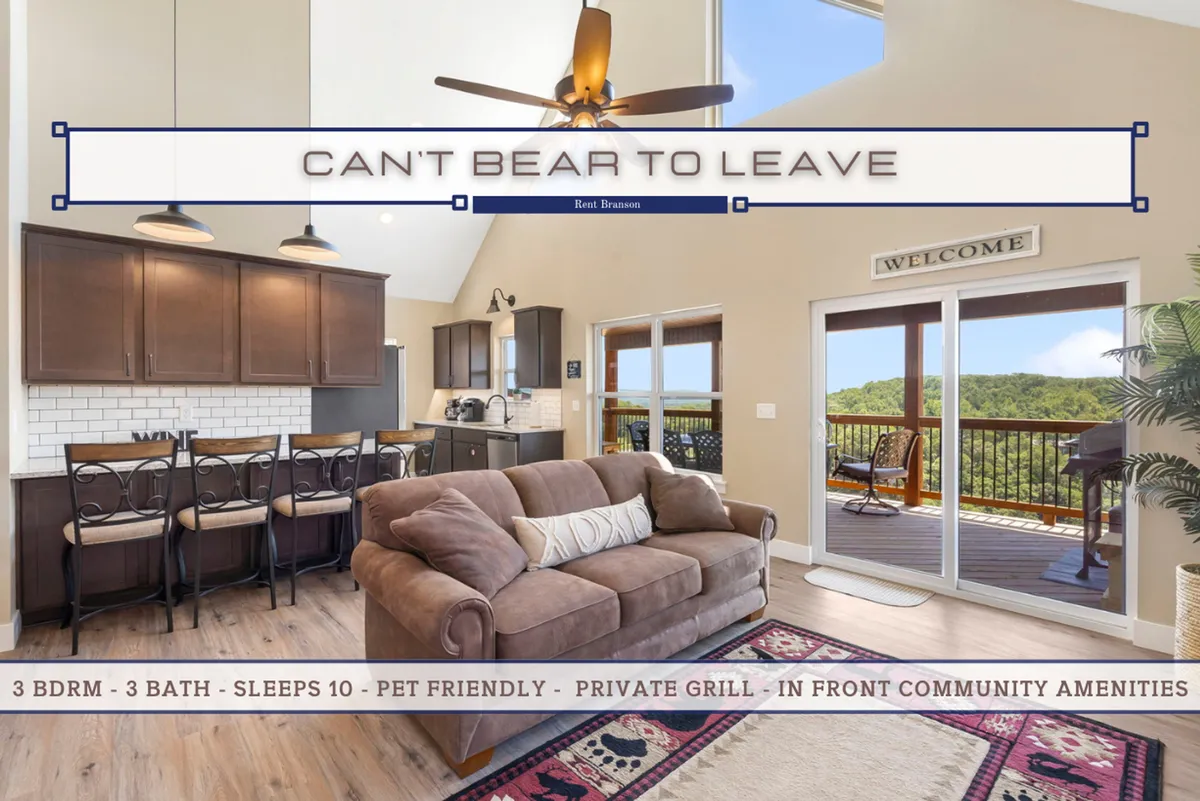 An expansive open-plan space featuring a central kitchen island, a plush sofa, and a spacious dining area, all framed by large windows letting in abundant natural light and showcasing views of the Ozarks. The area is styled for comfort and sociability, inviting group cooking, lounging, and togetherness.