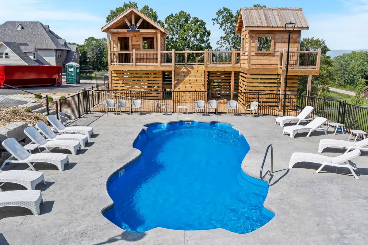 A lively outdoor setting featuring a swimming pool surrounded by wooden playhouses, lounge chairs, and children playing. The scene is vibrant and joyful, emphasizing family gatherings, playtime, and relaxing by the water.