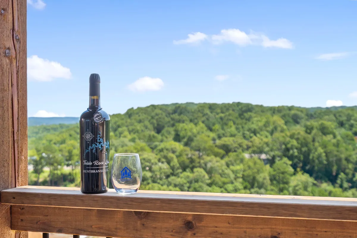 The image showcases a serene, mountainous landscape bathed in golden sunset light, viewed from a spacious outdoor deck. In the foreground, a bottle of wine and a glass rest invitingly on a table, suggesting leisurely relaxation while soaking in the panoramic wilderness views.
