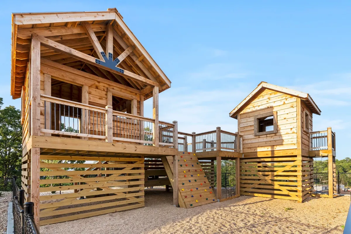 A whimsical treehouse-style playground with a playhouse structure, stairs, climbing wall, and lush surrounding greenery. Children are pictured playing and exploring, evoking a sense of imaginative adventure and joyful outdoor activity.