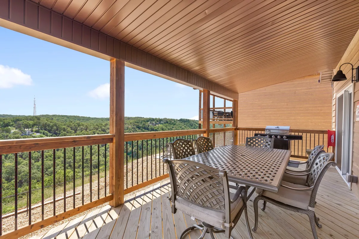 A spacious outdoor deck featuring a large dining table set for a meal, a high-quality grill ready for barbecuing, and comfortable seating overlooking a lush, forested view. The natural wood deck is bathed in soft daylight, inviting guests to gather and enjoy the outdoors.