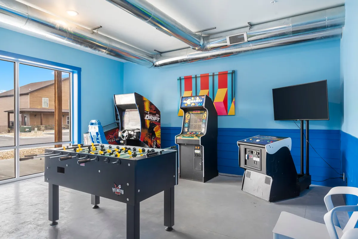 An energetic interior game room featuring a foosball table, several arcade machines, a television, and vibrant, colorful wall art. The space is designed for active entertainment and friendly competitions, appealing to both kids and adults.