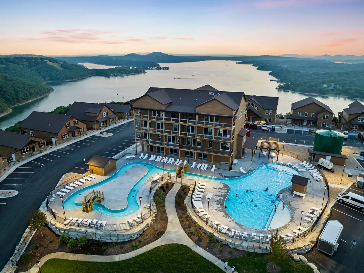A scenic exterior featuring sparkling outdoor swimming pools overlooking a lake, framed by the architecture of the resort and landscaped lounging areas. The view suggests both relaxation and recreational options, especially in warm weather.